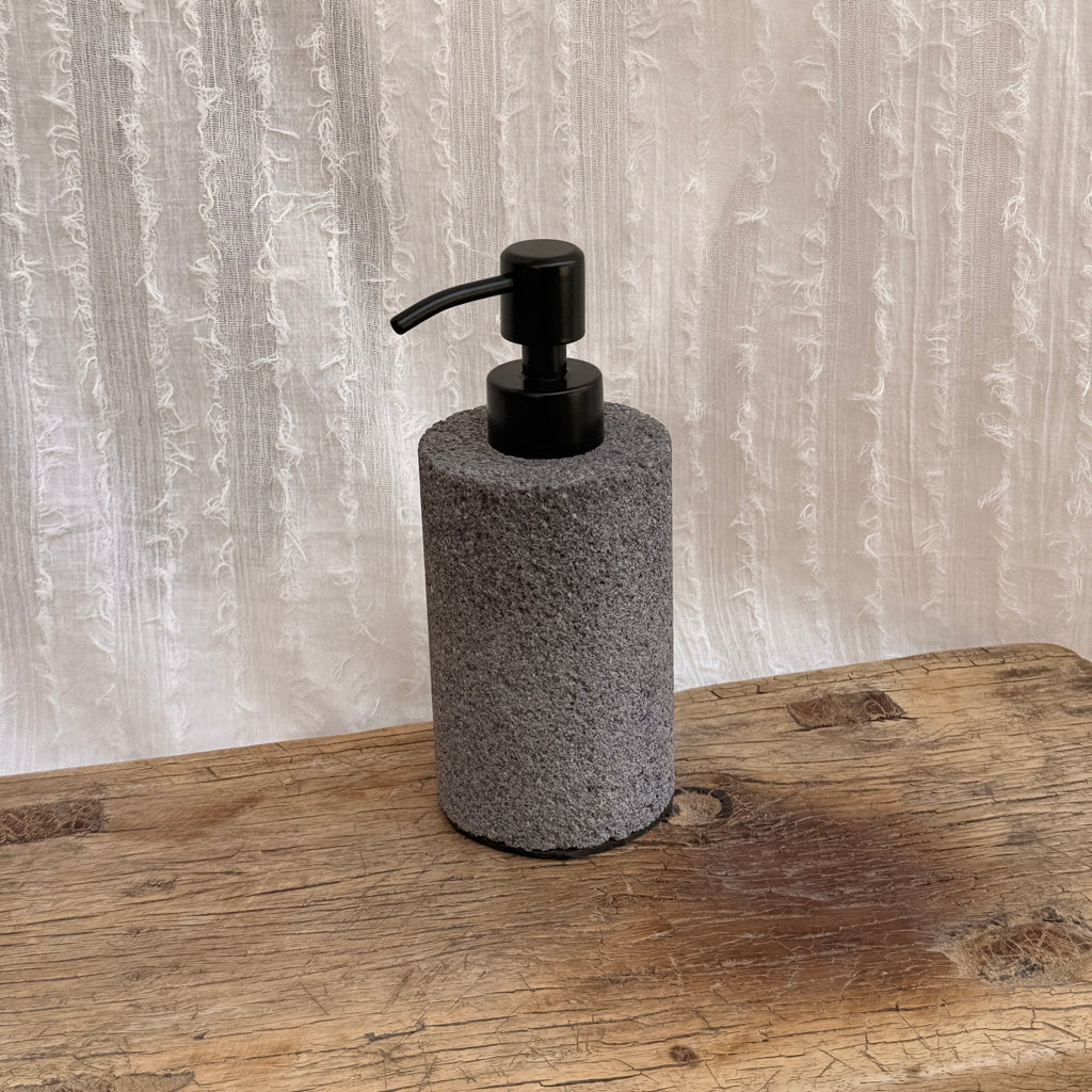 Lava rock soap dispenser with steel pump on vintage wooden surface with white textile background - wabi sabi bathroom
