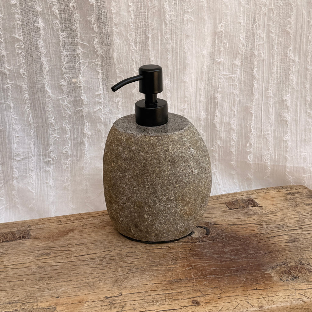 River stone soap dispenser with steel pump on vintage wooden surface with white textile background - wabi sabi bathroom